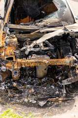 A car that caught fire is now a burnt shell, showing severe damage inside and out. The remnants of the engine and burnt materials are visible in a roadside setting