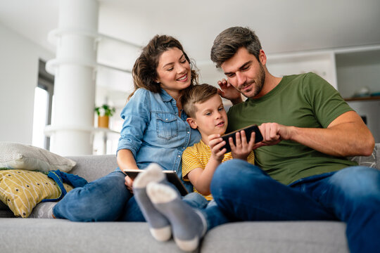 Happy parents with kid using smartphone at home together, playing, having fun with dog