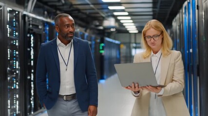 Two professionals inspect a server room, woman using laptop