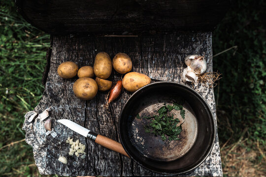 Pommes de terre, ail, &eacute;chalote et herbes fra&icirc;ches dispos&eacute;s sur une planche en bois avec un couteau et une casserole. Sc&egrave;ne rustique et naturelle de cuisine en plein air, &eacute;voquant la simplicit&eacute; 
