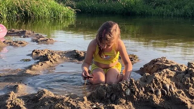 A girl in a yellow two-piece swimsuit plays in the sand on the river.