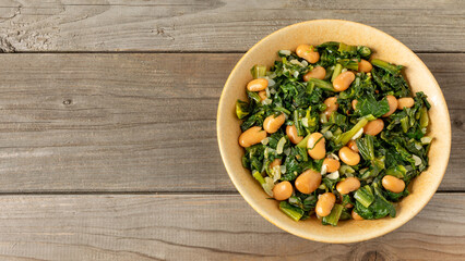 Top view of Italian chicory greens and white beans (Cicoria e Fagioli) with rosemary herb in a bowl on a rustic wooden background with space for text or logo.