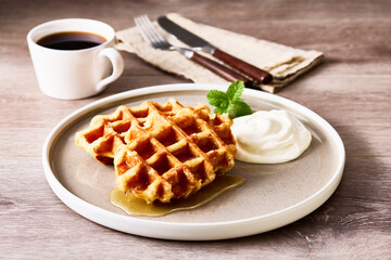焼きたてワッフルとホイップクリーム、メープルシロップを添えたティータイムのひととき。木のテーブルに置かれた白いプレートに、コーヒーと麻のナプキン、カトラリーを添えた温かみのあるブランチシーン