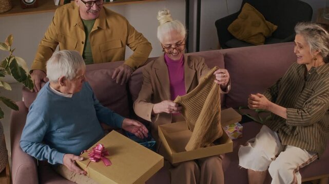 High angle shot of pleased elderly woman viewing presents she got for her 70th birthday from friends, she taking knitted vest and headscarf out of gift boxes while sitting on couch at home