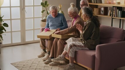 Happy elderly woman opening present taking brown vest out of box, she trying it on turning to friends while sitting on couch together during home birthday party - Powered by Adobe
