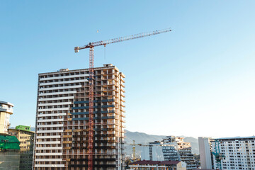 Fototapeta premium Modern high-rise building under construction with red tower crane against clear blue sky, surrounded by other contemporary buildings mountains in distance, concept of urban development architecture