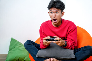Shocked Young Asian Male Playing Video Games With Controller And Sitting On Beanbag