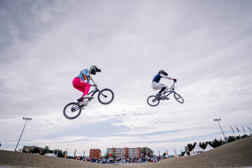 Fototapeta premium Valmiera, Latvia - July 12, 2025: BMX riders performing tricks in mid-air during competition with cloudy sky backdrop. BMX European championships