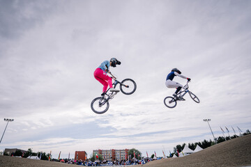 Fototapeta premium Valmiera, Latvia - July 12, 2025: BMX riders performing tricks in mid-air during competition with cloudy sky backdrop. BMX European championships