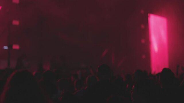 Silhouettes of people clapping and waving at a live music concert at an open-air festival. Fans applaud their favorite band. The stage is brightly lit.