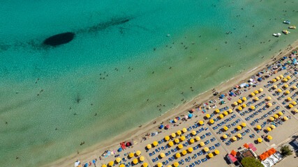 Aerial view of a beach resort on a sandy beach. The sea is crystal clear and emerald green. The beach club has yellow umbrellas, blue loungers, and vacationers sunbathing.