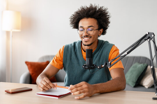 Smiling, happy African American man, podcaster using microphone talking taking notes in home studio - Powered by Adobe