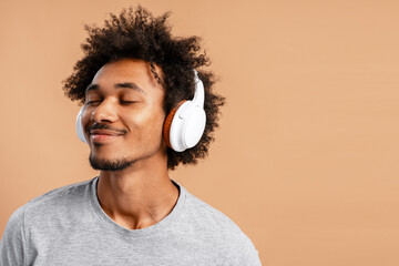 African American man listening music in headphones with closed eyes isolated on beige background