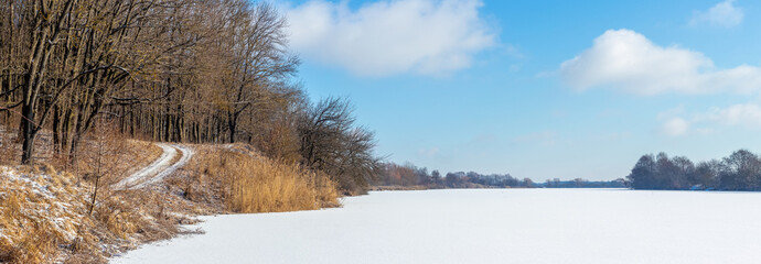 Panoramic winter landscape with a frozen lake or river covered with snow, blue sky and a forest on...