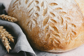  Close up a loaf of artisanal bread rests on a gray napkin, showcasing intricate leaf patterns on its crust. Surrounding it are golden wheat stalks, highlighting its rustic appeal.