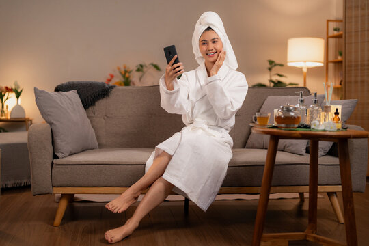 Asian adult woman home spa pamper session using smartphone selfie video call relaxing cozy living room beauty routine after bath wellness
