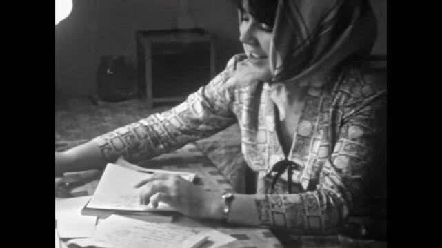 Young woman writes in notebook, making homework notes at table in cozy home. Close up, Girl looking to camera, shy smile. Focused student studying. Old film. Archival retro 1960s. Vintage archive