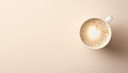 Overhead view of a single cup of cappuccino.  A heart latte art design atop the light-brown coffee drink in a white mug.  Pale beige background