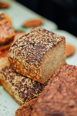 Close-up of rustic whole grain bread topped with toasted seeds, showcasing a crisp crust and dense texture. Loaves are stacked on a light-colored surface