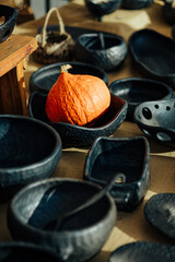A vibrant orange pumpkin rests inside a textured black ceramic bowl, surrounded by other handcrafted pottery pieces on a display table at a market