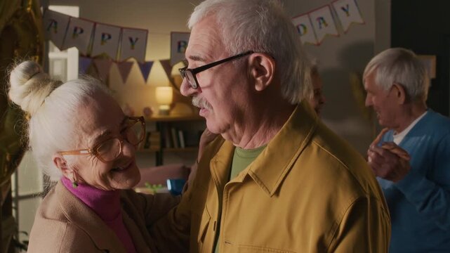 Happy 70-year-old lady dancing and talking with elderly male friend at home birthday party, both wearing glasses and having grey hair