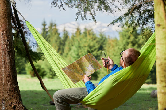 Man traveler in glasses studies map while holding compass, planning outdoor adventure or hiking route, relaxes in green hammock in forested area. Trees with distant mountains on blurred background.