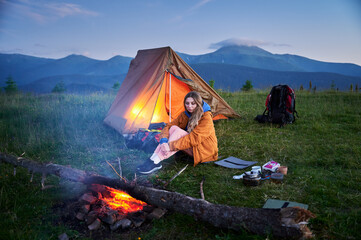 Young woman tourist sits beside campfire near illuminated tent, against backdrop of majestic...