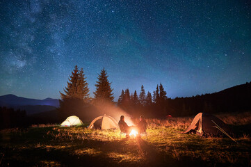 Two people tourists relax in chairs by campfire, with illuminated tent nearby, under breathtaking...