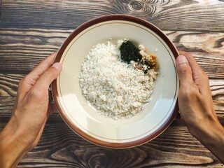 Hand holding a plate of traditional Indonesian white corn rice, served with anchovies, boiled fennel leaves, and fresh spicy sambal.