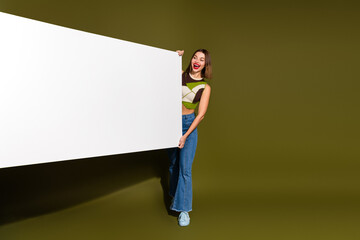 Young woman with stylish outfit holds blank white banner over solid khaki background, smiling...