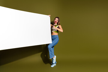 Cheerful young woman holding a large blank banner on a khaki background, expressing joy and stylish...