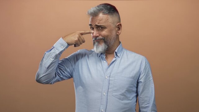Man pointing finger to temple in studio with peach backdrop and light blue shirt, gray beard and raised eyebrow; wry humor skepticism.