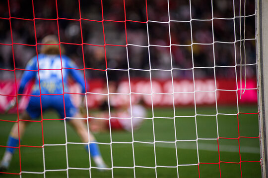 Two-tone goal net in focus with blurred goalkeeper and striker beyond. Dynamic football moment with selective focus and game intensity.