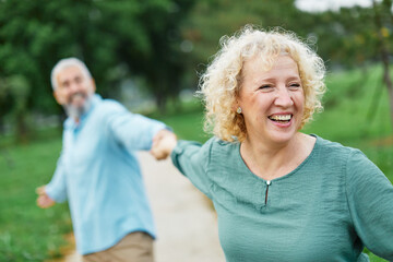 Happy active mid aged adult couple having fun walking running holding hands and bonding in park or...