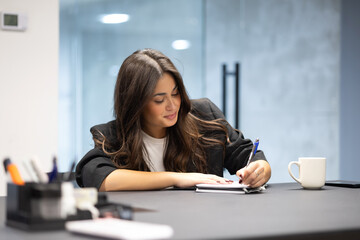 A focused young businesswoman sits at a desk in an office, writing in a notebook