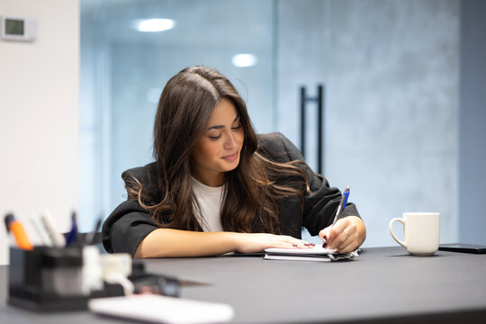 A focused young businesswoman sits at a desk in an office, writing in a notebook - Powered by Adobe