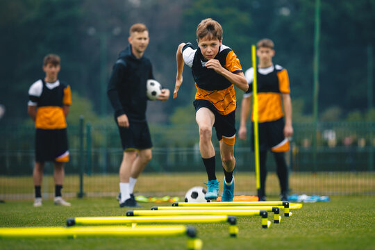 Teenage Boy at Training Drill With Young Soccer Coach. Youth Football Player Jumping Over Row of Hurdles and Running Fast at Sports Practice Field. Sports Strength and Agility Training For Footballers
