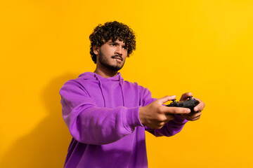 Cheerful man playing video games with focused expression against a vibrant yellow background wearing a purple hoodie