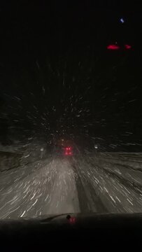 Vertical POV video of driving through a snowy night storm, seen from inside a car. Snow hits the windshield as headlights blur in traffic