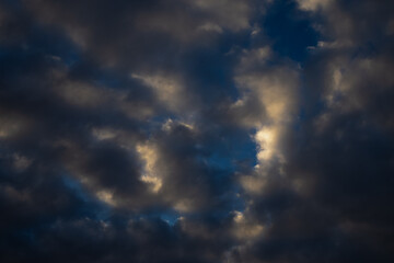 Dark evening sky with dramatic cloud formations illuminated by faint light, showing contrast between shadow and color layers within natural atmospheric depth and moody weather texture