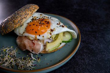 Modern breakfast with bagel sandwich, fried egg, prosciutto and avocado, topped with sesame, poppy seeds, served on ceramic plate,  side view on black background.