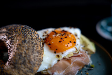 Breakfast sandwich with fried egg, prosciutto and avocado, topped with sesame and poppy seeds, served on matte teal dish, side view on black background.