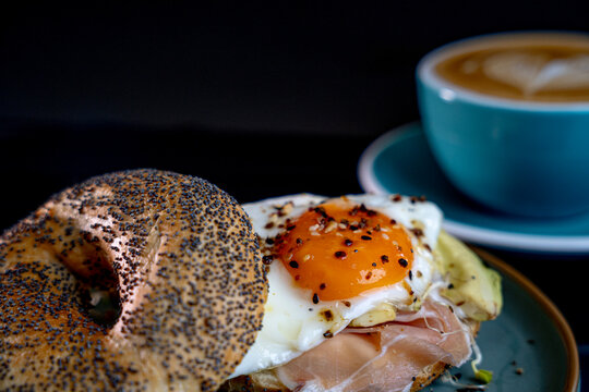 Gourmet breakfast with bagel sandwich, fried egg, prosciutto and avocado, topped with sesame, poppy seeds, served on ceramic plate, and cup of cappuccino with latte art, side view on black background - Powered by Adobe