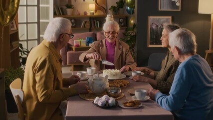 Stylish senior lady cutting birthday cake and sharing it with friends during tea party at festive table in living room