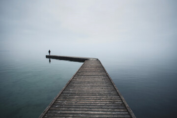 Fototapeta premium Long pier on the sea with a man backlit gazing into infinity, illuminated by the evening lights