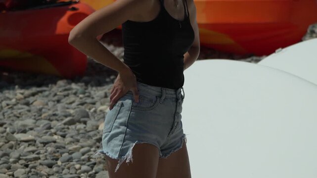 Woman, shorts, beach woman in denim shorts standing on a rocky beach with kayaks in background, summer day.