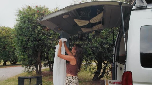 Young adult woman hangs a towel outside her camper van in a natural park setting, enjoying a warm and sunny day amidst trees.