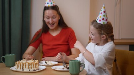 Loving mother sharing birthday celebration with daughter, wearing festive party hats and enjoying delicious cake while creating heartwarming family memories at home - Powered by Adobe