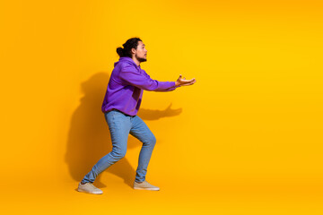 Young man gesturing with outstretched hands on a bright yellow background wearing a purple hoodie and casual jeans