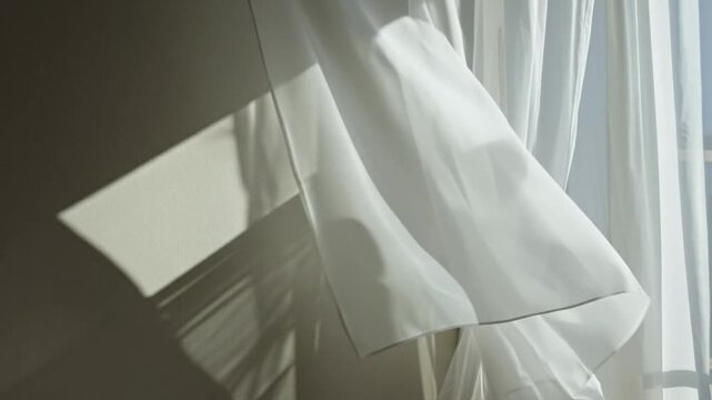 White sheer curtains gently billow in the sunlight, casting long, peaceful shadows on an indoor wall.